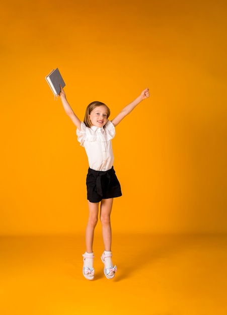 Gelukkig schoolmeisje in een uniform springt met een blauw notitieboekje op een gele achtergrond met een kopie van de ruimte. Terug naar school