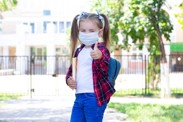 Gelukkig schoolmeisje met een beschermend masker met een rugzak en een leerboek in haar handen. in een wit T-shirt en een geruit overhemd