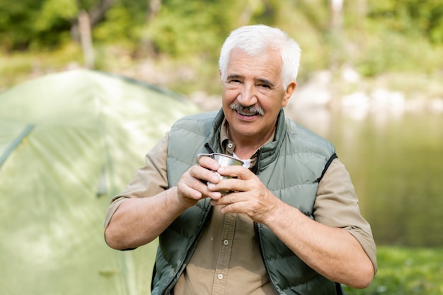 Gelukkig senior actieve man met warme drank op zoek naar jou terwijl je geniet van zijn reis in het bos op zomerweekend