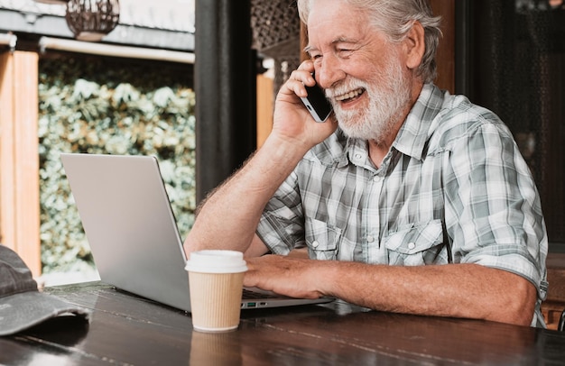 Gelukkig senior bebaarde man browsen op internet door laptop Knappe oudere man zittend buiten op een houten tafel praten op mobiele telefoon dicht bij een afhaalmaaltijden koffie