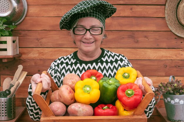 Foto gelukkig senior boer houdt een houten mand vol met vers geplukte paprika's en aardappelen uit de tuin.