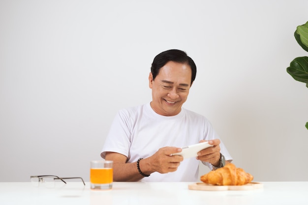 Gelukkig senior koppel dat thuis een croissantontbijt heeft en sinaasappelsap drinkt, met behulp van smartphone.