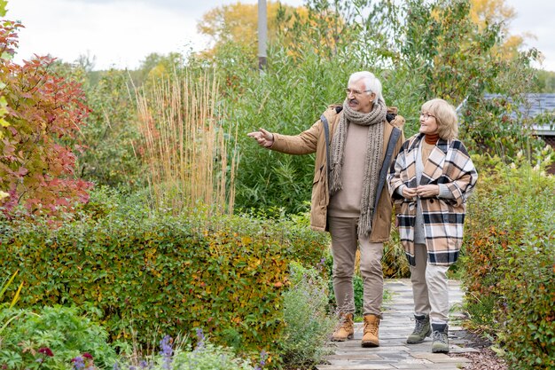 Gelukkig senior koppel in warme vrijetijdskleding die over een smalle weg tussen struiken en bomen beweegt terwijl ze over planten in hun tuin praten