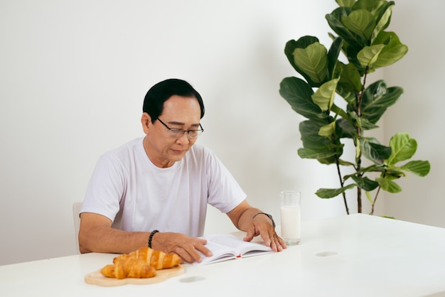 Gelukkig senior paar met croissant en melk ontbijt thuis, leesboek.