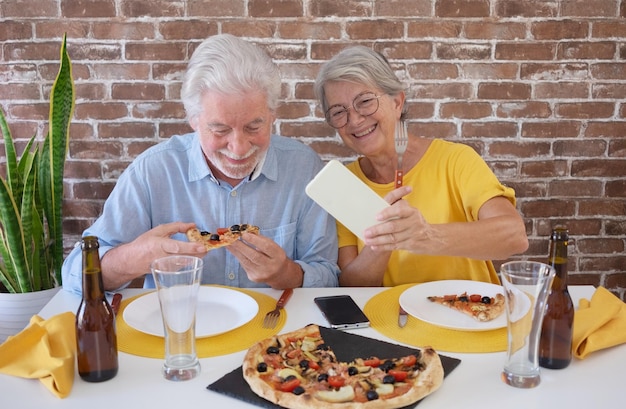 Gelukkig senior paar plezier samen met pizza en bier oudere vrouw en man lachen om thuis tafel te zitten met behulp van telefoon voor een selfie