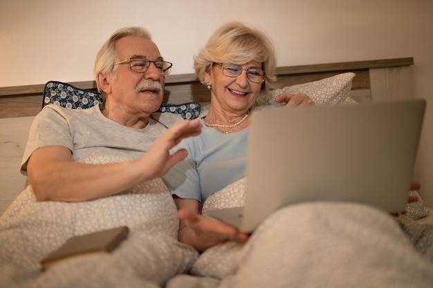 Gelukkig senior paar zwaaien naar iemand tijdens het maken van video-oproep via laptop in slaapkamer