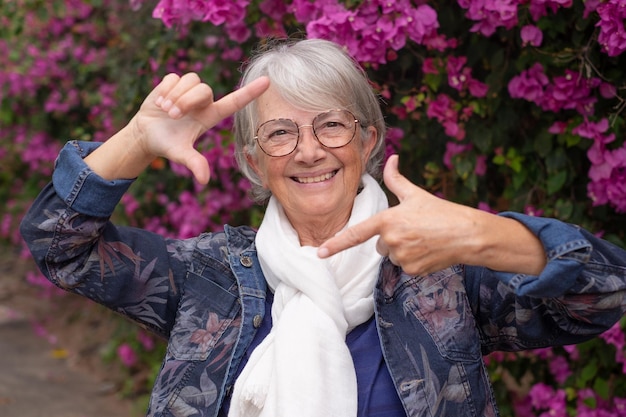 Gelukkig senior vrouw in het park in de buurt van roze bougainvillea kijkt naar camera maken fotolijstjes met handen Lachende oudere dame met bril en wit haar genietend van haar vrije tijd en pensioen buitenshuis