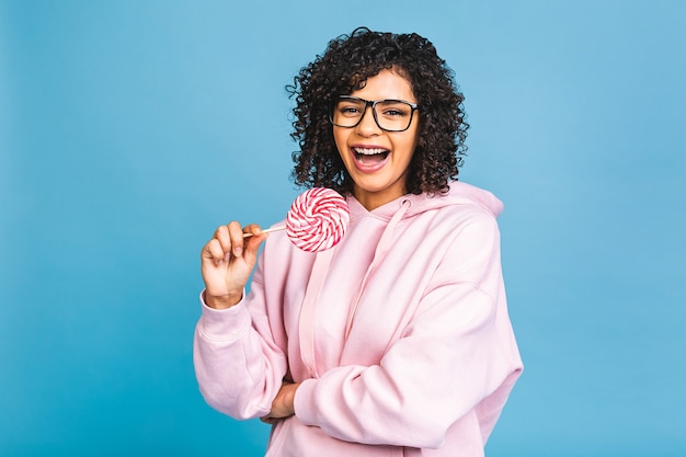 Gelukkig sexy Amerikaans afromeisje dat lolly eet. Schoonheid glamour model vrouw met zoete kleurrijke lolly snoep, geïsoleerd op blauwe achtergrond. Snoepgoed.