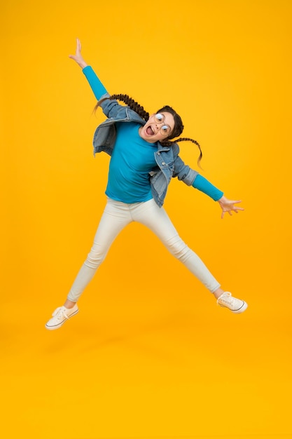 Gelukkig springend tienermeisje met gevlochten haar draagt een bril voor het gezichtsvermogen op gele achtergrond terug naar school