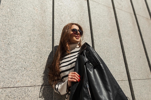 Gelukkig stedelijk modieus meisje met zonnebril in een gestreepte modetrui met een stijlvol leren zwart jasje staat op een zonnige dag bij de muur in de stad Straatmode en beauty