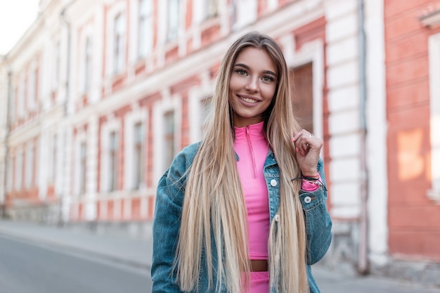 Gelukkig stedelijke jonge vrouw met positieve glimlach met lang blond haar in een trendy jeansjasje in een stijlvolle roze top vormt in de buurt van een vintage wit gebouw in de straat. Vrij vrolijk meisje loopt in de stad.