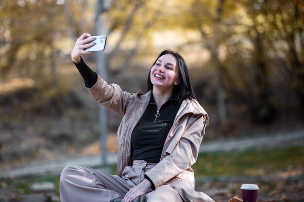 Gelukkig stijlvol vrouwenportret in de herfst met trenchcoat, ze neemt een selfie