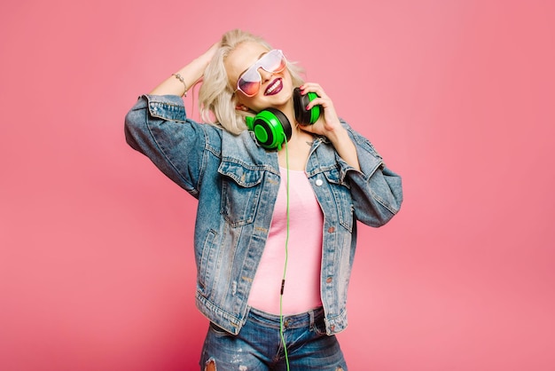 Gelukkig stijlvolle blonde vrouw in roze bril met grote koptelefoon muziek luisteren en dansen op gekleurde achtergrond