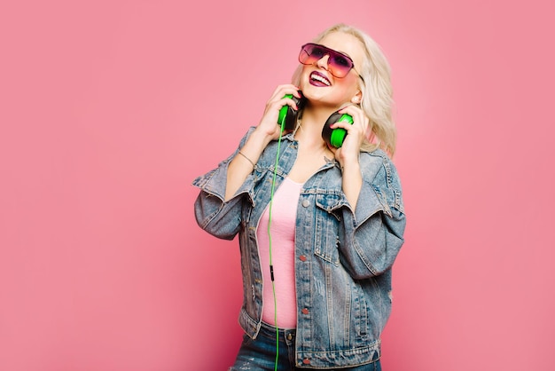 Gelukkig stijlvolle blonde vrouw in roze bril met grote koptelefoon muziek luisteren en dansen op gekleurde achtergrond