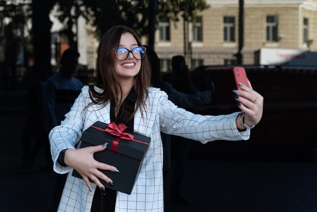 Gelukkig stijlvolle brunette met een cadeau in haar handen maakt een selfie op een smartphone.
