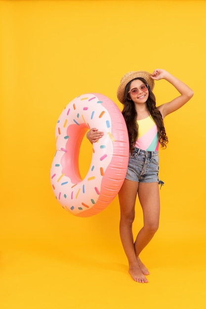 Gelukkig strand kind met krullend haar in stro hoed en zonnebril met donut opblaasbare ring voor pool party plezier op zomervakantie op gele achtergrond zomervakantie