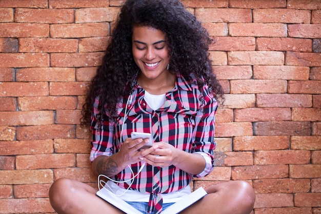 Gelukkig student vrouw met afro kapsel zitten met bakstenen muur achtergrond met behulp van mobiele telefoon en glimlachen.