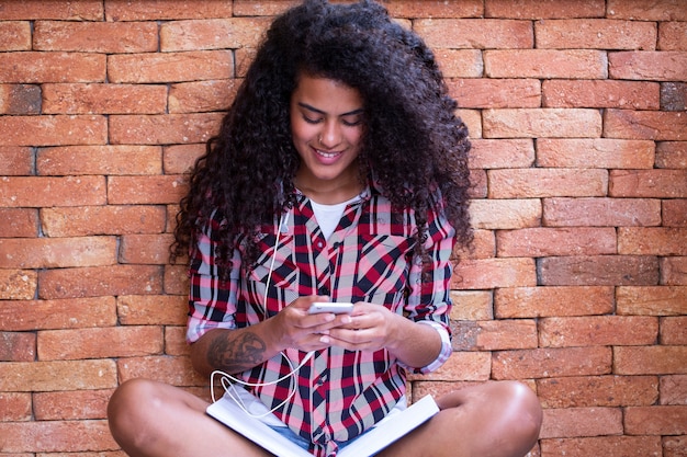 Gelukkig student vrouw met afro kapsel zitten met bakstenen muur achtergrond met behulp van mobiele telefoon en glimlachen.