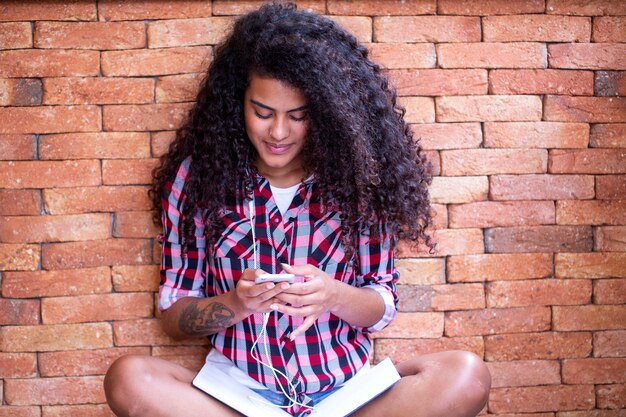 Gelukkig student vrouw met afro kapsel zitten met bakstenen muur achtergrond met behulp van mobiele telefoon en glimlachen.