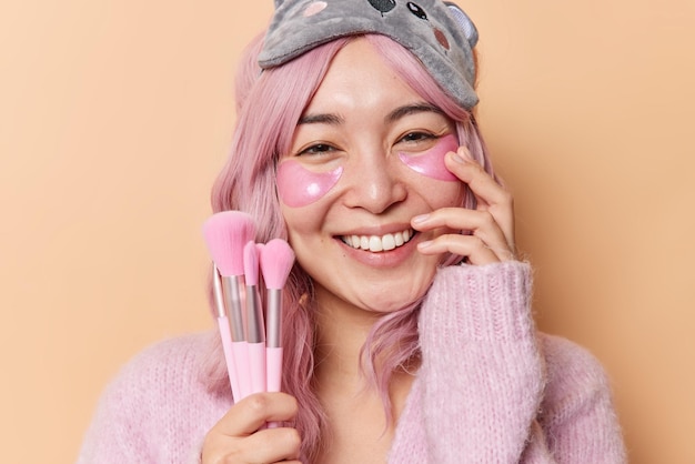 Gelukkig tedere Aziatische vrouw met roze haar houdt cosmetische borstels past hydrogel patches voor huidbehandeling draagt zachte trui en slaapmasker glimlacht graag geïsoleerd over beige studio achtergrond