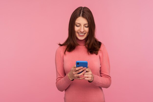 Gelukkig tevreden brunette vrouw chatten op smartphone met brede glimlach op het gezicht, winkelen, tickets boeken, eten online bezorgen. Indoor studio-opname geïsoleerd op roze background