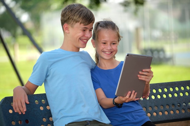 Gelukkig tiener kinderen meisje en jongen kijken naar scherm van digitale tablet lezen studeren of gamen zittend op een bankje buiten op zonnige zomerdag