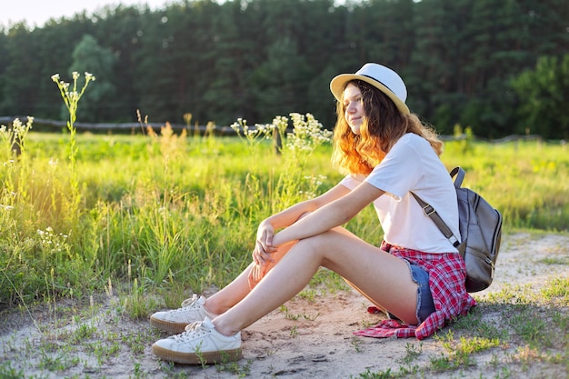 Gelukkig tienermeisje in hoed met rugzak zittend op landweg, genietend van zomervakantie, pittoreske natuur, kopieer ruimte