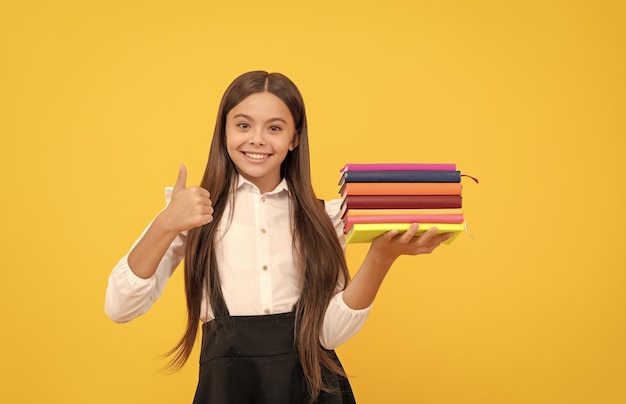 Gelukkig tienermeisje in schooluniform houdt boekenstapel vast en toont duim omhoog gebaar excellentie