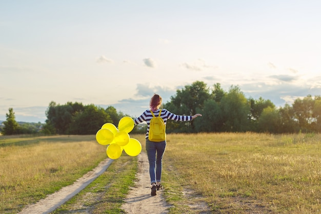 Gelukkig tienermeisje met gele ballonnen en rugzak rennen en springen langs de landweg in de zomerweide. vrijheid, leven, vreugde, vakantieconcept