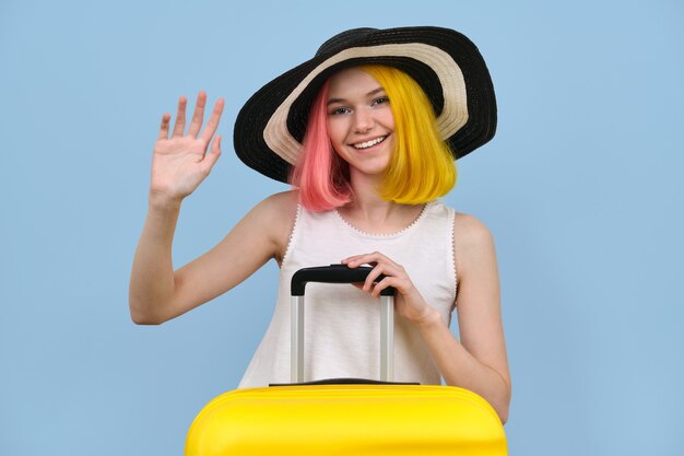 Gelukkig tienermeisje met koffer in hoed zwaait met zijn hand op blauw gekleurde achtergrond. Vakantie, hallo, doei, vakantie, jeugdconcept