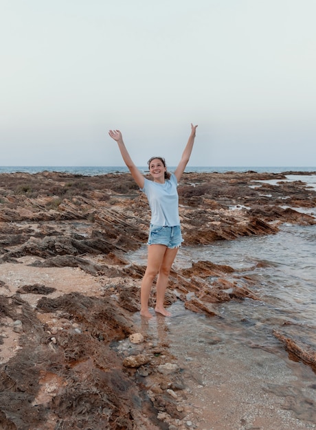 Gelukkig tienermeisje staande op de klif aan zee bij zonsondergang met een lichtblauw t-shirt, een spijkerbroek en haar handen omhoog. t-shirtmodel