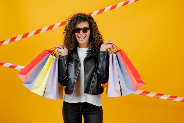 Gelukkig trendy zwart meisje met kleurrijke die het winkelen zakken over geel met signaalband wordt geïsoleerd