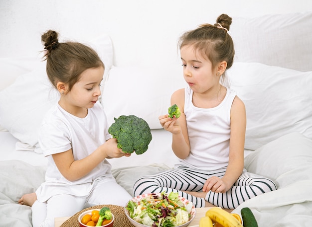 Gelukkig twee schattige meisjes fruit en groenten eten in de slaapkamer op het bed. Gezonde voeding voor kinderen en tieners.