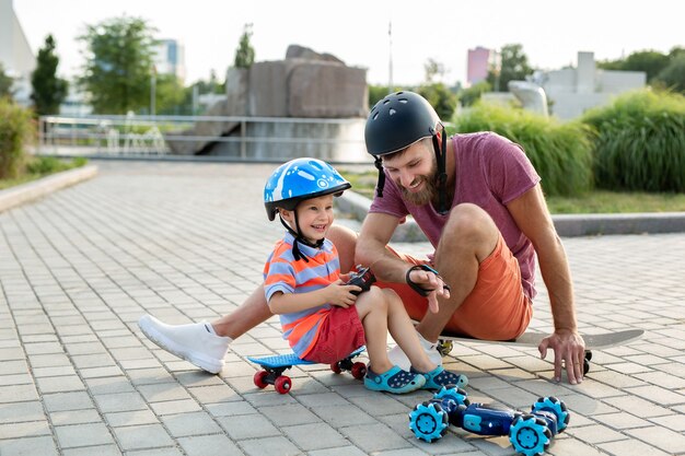 Gelukkig vader en zoon in helmen spelen in het park met een robotauto die wordt bestuurd door een handschoen zittend op skateboards.