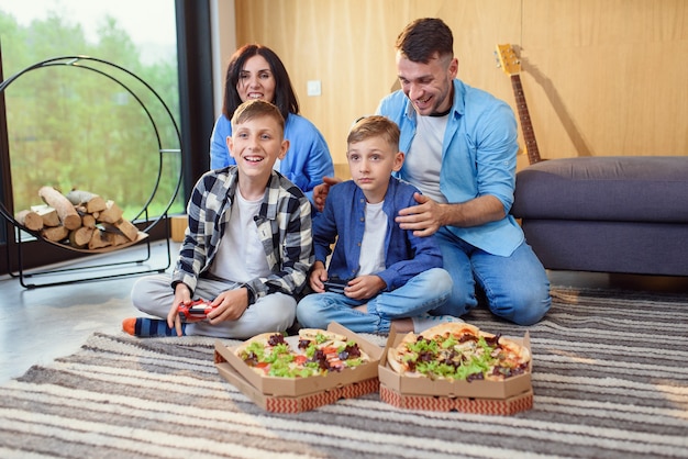 Gelukkig vader moeder en twee zonen zittend op de vloer spelen van videogames met gamepads en smakelijke pizza eten