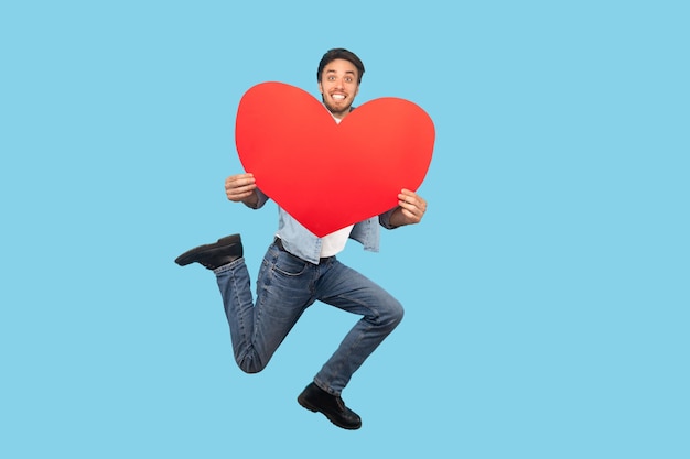 gelukkig verliefd man springt en houdt grote rode hartvorm vast en kijkt met liefde en brede glimlach. indoor studio-opname geïsoleerd op blauwe achtergrond