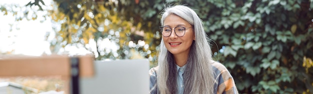 Gelukkig volwassen aziatische vrouw met bril en aardbeiendessert voor laptop aan tafel buiten