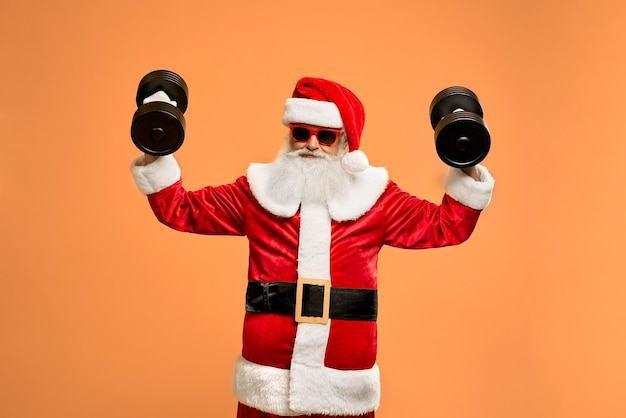 Gelukkig volwassen kerstman met grijsharige baard in rode zonnebril en kerstpak oefenen met twee zware zwarte halters in studio. Lichaamsbeweging en gezonde levensstijl concept.