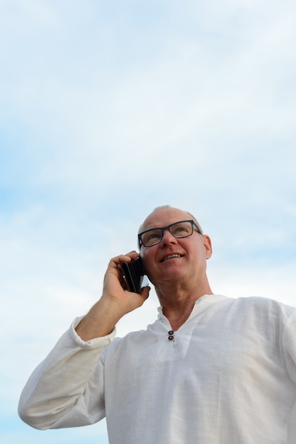 Gelukkig volwassen knappe toeristische man praten aan de telefoon tegen uitzicht op het strand buitenshuis