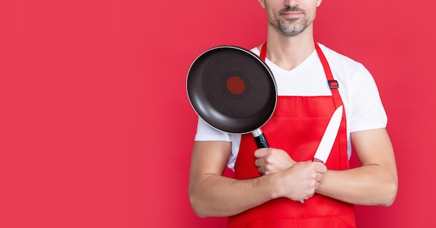 Gelukkig volwassen man chef-kok in schort met koekenpan en mes op rode achtergrond