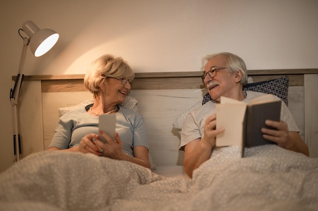 Gelukkig volwassen stel dat 's avonds met elkaar praat terwijl ze in de slaapkamer liggen Man leest een boek terwijl vrouw smartphone gebruikt
