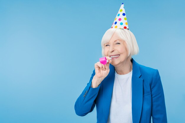 Gelukkig volwassen vrouw in feestmuts maken lawaai geïsoleerd op blauwe achtergrond