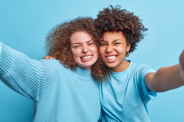 Gelukkig vriendelijk gemengd ras jonge vrouwen omarmen ans pose voor selfie glimlach met tanden hebben cheeful uitdrukkingen terloops gekleed geïsoleerd over blauwe muur. Mensen emoties leuk vriendschap concept