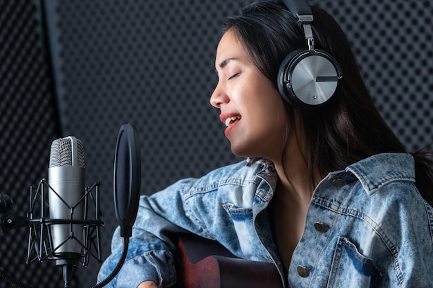 Gelukkig vrolijk mooi lachend portret van een jonge Aziatische vrouw zanger die een koptelefoon draagt met een gitaar die een nummer van de microfoon opneemt in een professionele studio