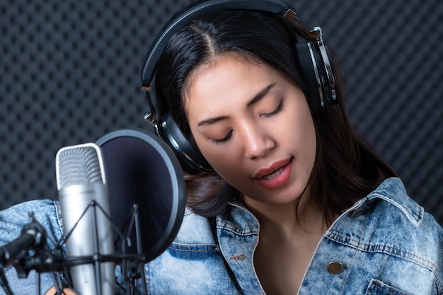 Gelukkig vrolijk mooi lachend portret van een jonge Aziatische vrouw zanger die een koptelefoon draagt met een gitaar die een nummer van de microfoon opneemt in een professionele studio