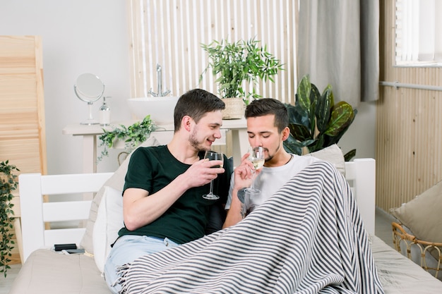 Gelukkig vrolijk paar dat op bed met glazen wijn thuis ligt. Homopaar die en in bed koesteren kussen. Homopaar Love Home Concept