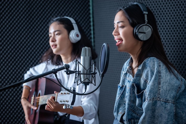 Gelukkig vrolijk vrij lachend van portret twee jonge Aziatische vrouw zanger die een koptelefoon draagt met een gitaar die een lied voor microfoon opneemt in een professionele studio