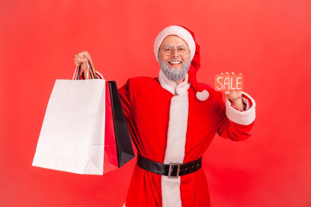 Gelukkig vrolijke kerstman houden en tonen verkoop kaart en kleurrijke papieren zakken kijken camera met brede glimlach tevreden met vakantie kortingen Indoor studio opname geïsoleerd op rode achtergrond