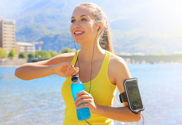 Gelukkig vrolijke vrouwelijke atleet met armband voor slimme telefoon power drinkfles openen na dagelijkse training en buitenshuis wegkijken.