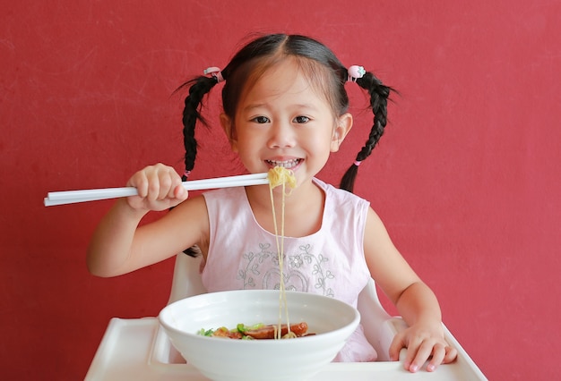 Gelukkig weinig Aziatisch meisje die Einoedel op hoge stoel eten tegen rode muurachtergrond.